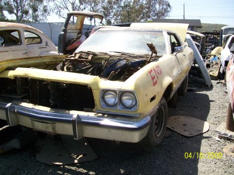 1972 Ford Ranchero - A faded yellow 1970s Ford coupe sits in a junkyard with its hood removed, exposing the engine bay, missing headlight trim, and rusted body…