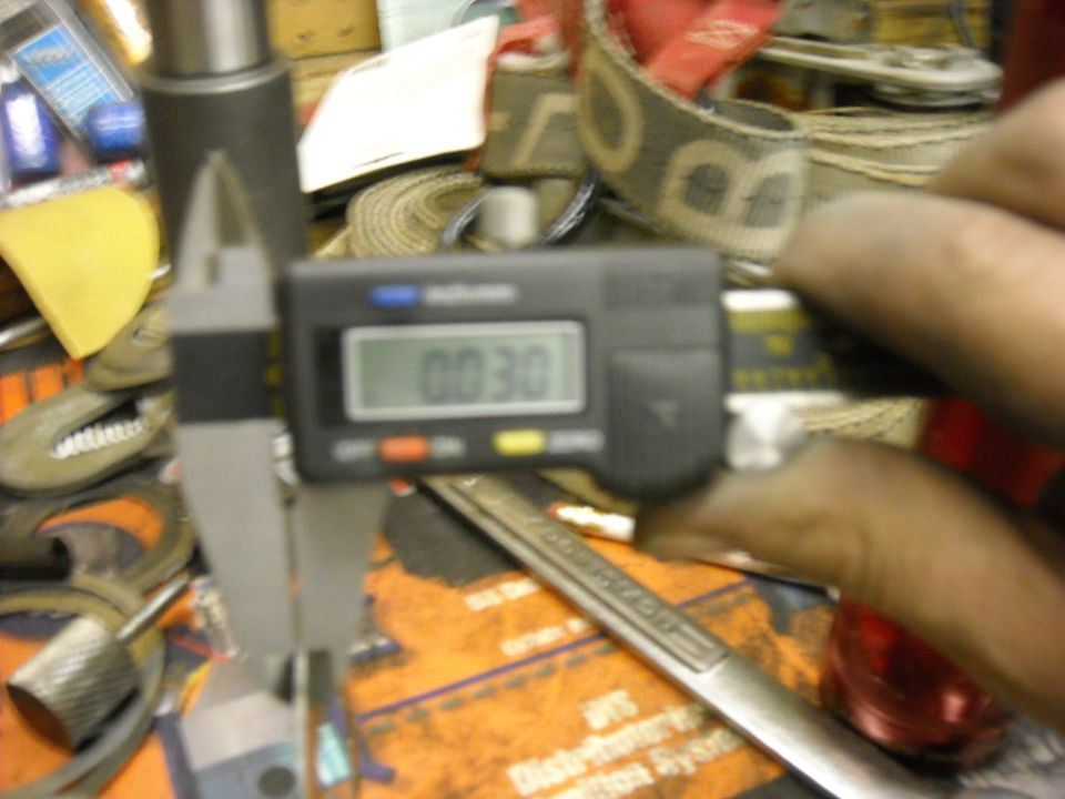 Digital caliper showing 0.030 measurement, held by a hand. Background has tools and straps
