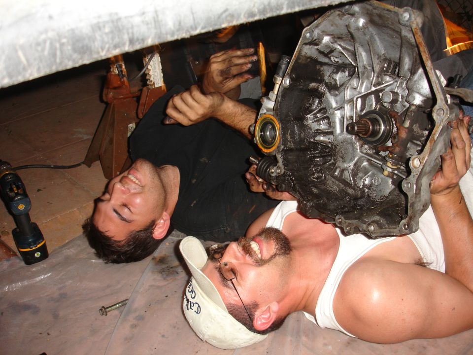 Two men lying on the ground under a vehicle, one holding a transmission