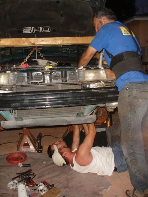 1995 Ford Taurus - Two mechanics are working on the engine of a 1995 Ford Taurus SHO, which is supported by jack stands.