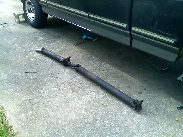 Driveshaft removed from a dark-colored truck, lying on concrete. Truck is on blocks