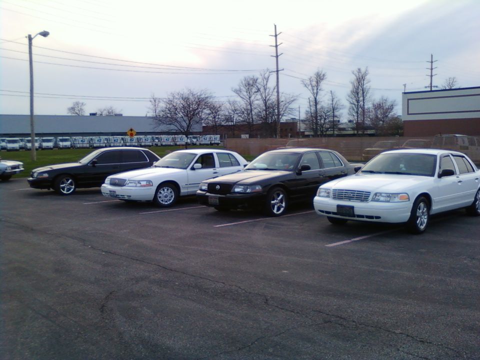 Four sedans parked in a lot, white Crown Victoria-style car at right, overcast sky