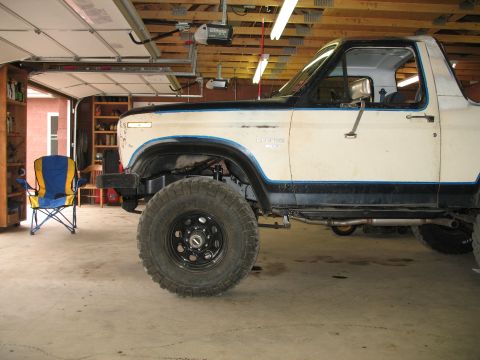 1985 Ford Bronco - A white Ford Bronco with a blue stripe sits in a garage next to a folding chair.