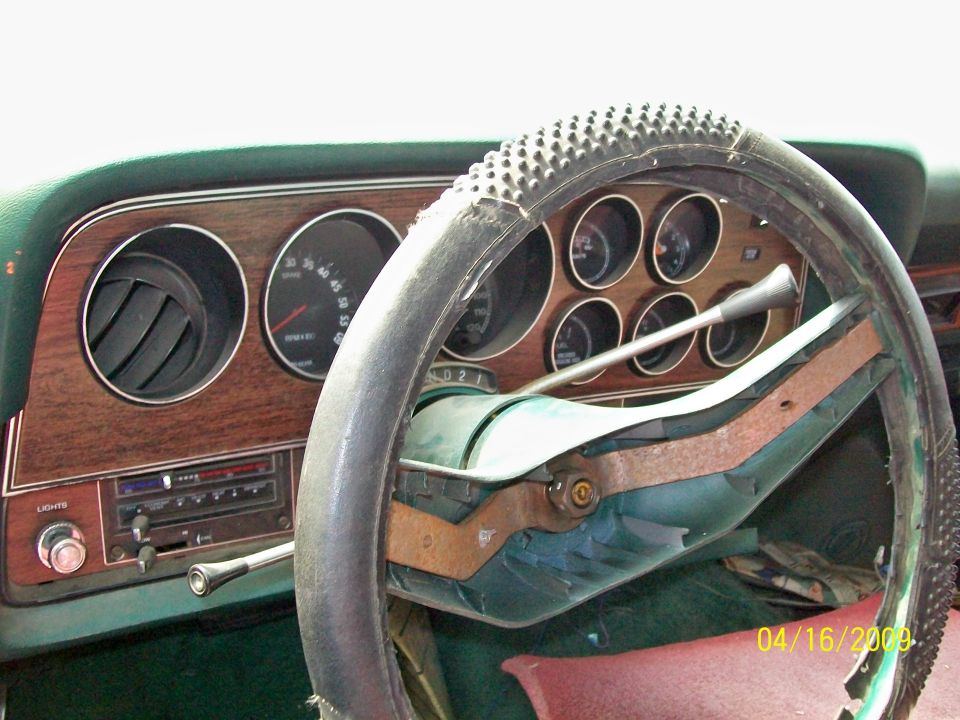 Dashboard of 1972 Ford Ranchero seen through worn steering wheel with exposed rusty spokes and woodgrain gauge panel.