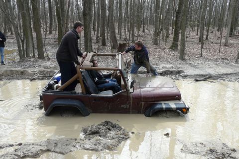 1995 Ford Bronco - A maroon Jeep is submerged in a muddy forest clearing, with several people attempting to free it.