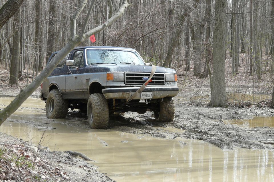 Ford Bronco with lifted suspension driving through a muddy forest trail. License plate reads 292W24