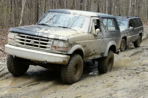 1995 Ford Bronco - A muddy 1995 Ford Bronco navigates an off-road trail, followed closely by a Jeep Grand Cherokee.