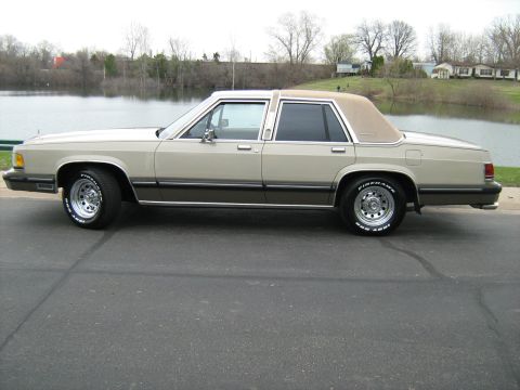 1988 Mercury Grand Marquis - A two-tone 1988 Mercury Grand Marquis sedan is parked on a road with a lake and houses in the background.