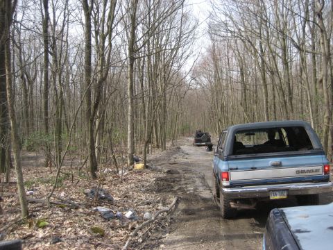 1991 Ford Bronco - A blue Chevrolet K5 Blazer navigates a wooded dirt road, with another vehicle visible ahead.