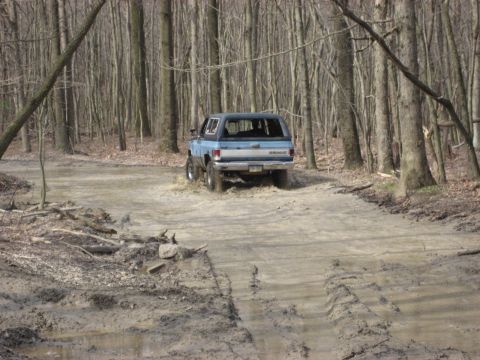 1991 Ford Bronco - A blue SUV navigates a muddy path surrounded by trees.