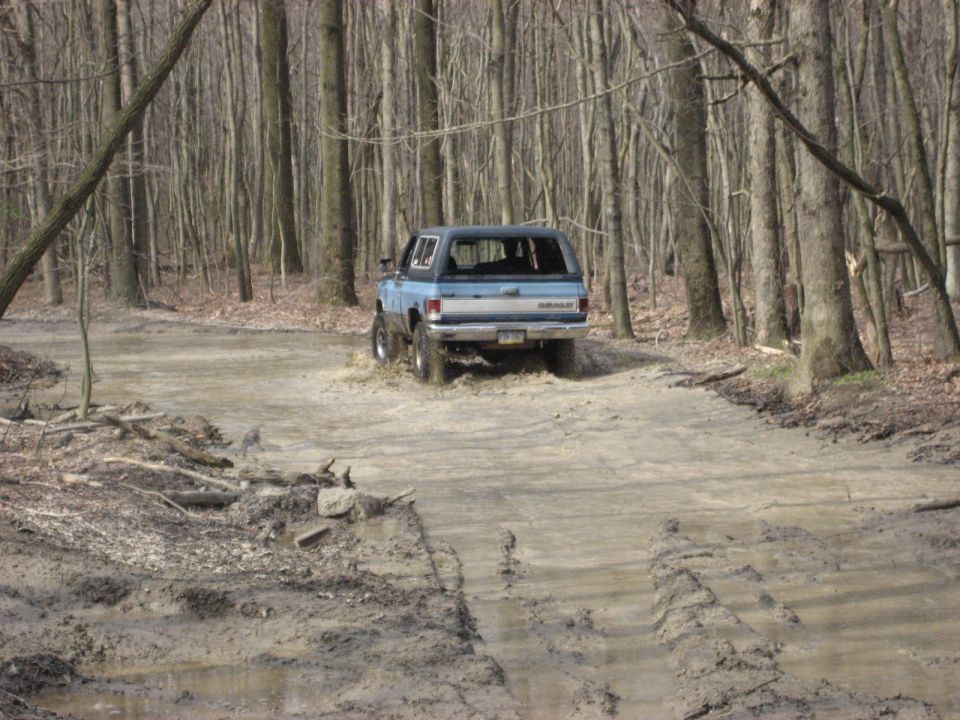 Blue and white square-body Chevy Blazer driving through a flooded muddy wooded trail
