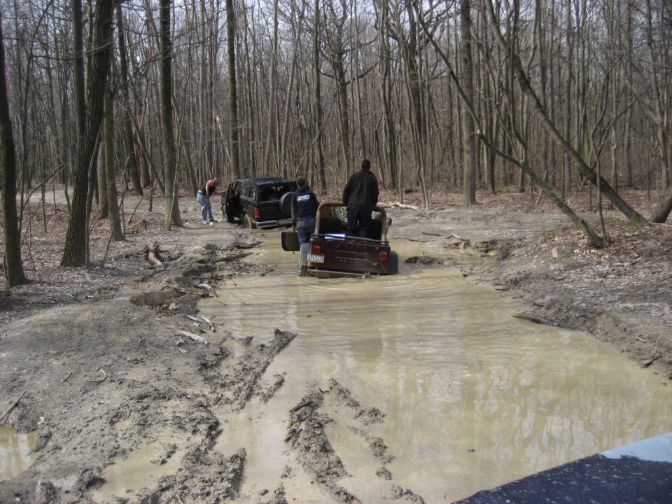 Brown SUV and black SUV stuck in mud, trees and dirt road