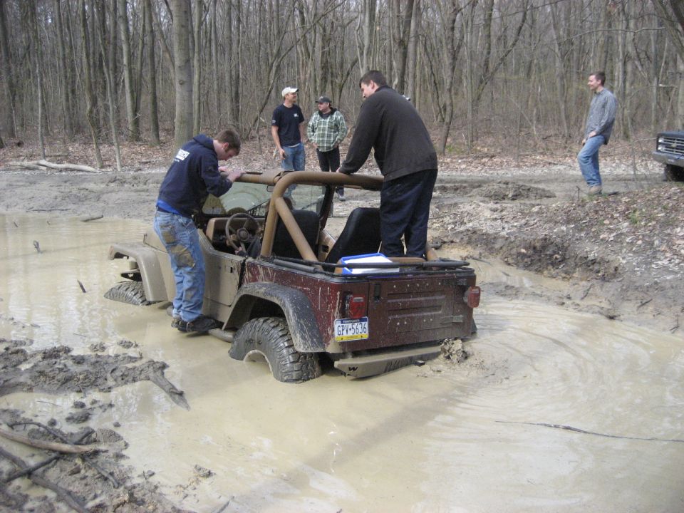 Jeep Wrangler stuck in mud pit with several people attempting to help; another vehicle visible in background