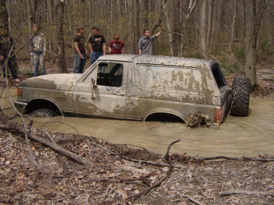 Mud-covered white Ford Bronco sunk in a muddy trail pond, side view
