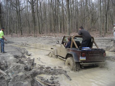 1991 Ford Bronco - A tan Jeep Wrangler is stuck in the mud while being pulled by a chain in a wooded area.