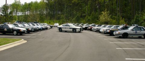 1993 Ford Mustang - Multiple 1993 Ford Mustang police cars are parked in a lot with trees in the background.