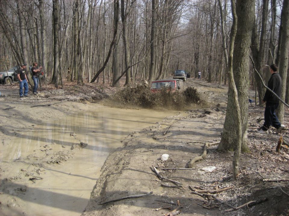 Several off-road vehicles navigate a muddy track in a wooded area; one red vehicle splashes through a deep puddle