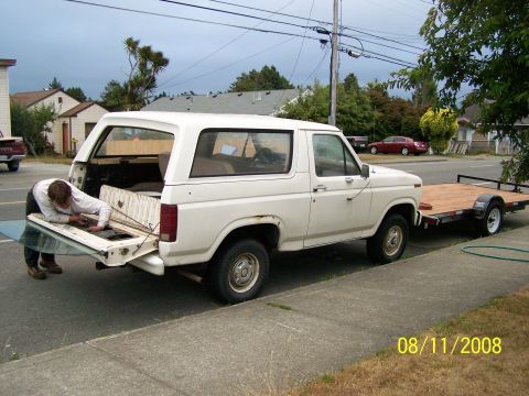 1982 Ford Bronco - A person works on the open tailgate of a white 1982 Ford Bronco, which is hitched to a wooden trailer.