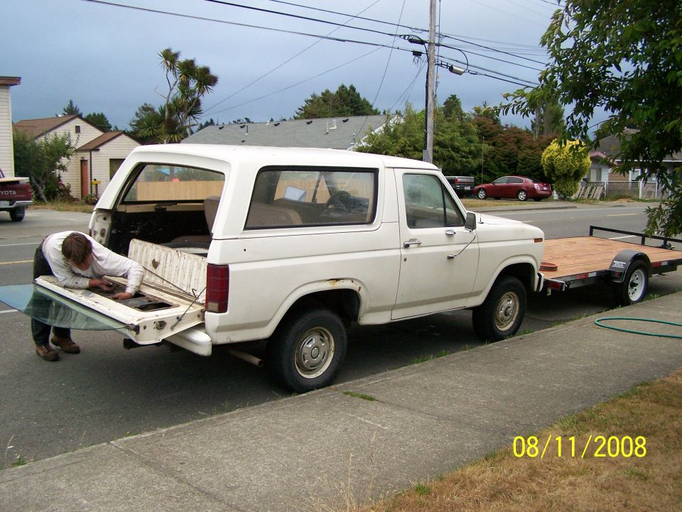 White 1982 Ford Bronco side view, tailgate open, person working inside, towing trailer