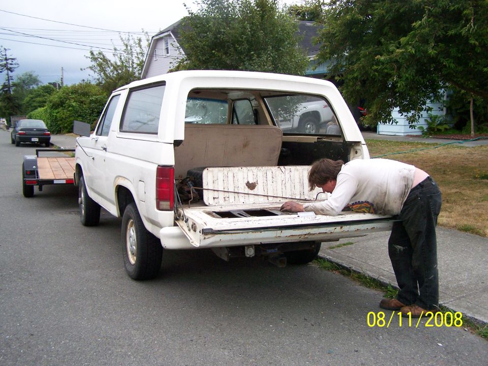White 1980s Ford Bronco towing a trailer, viewed from the rear with a person leaning into the open tailgate