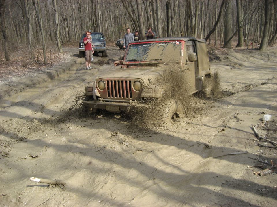 Mud-covered Jeep Wrangler with red roll bar splashing through a deep mud pit in the woods