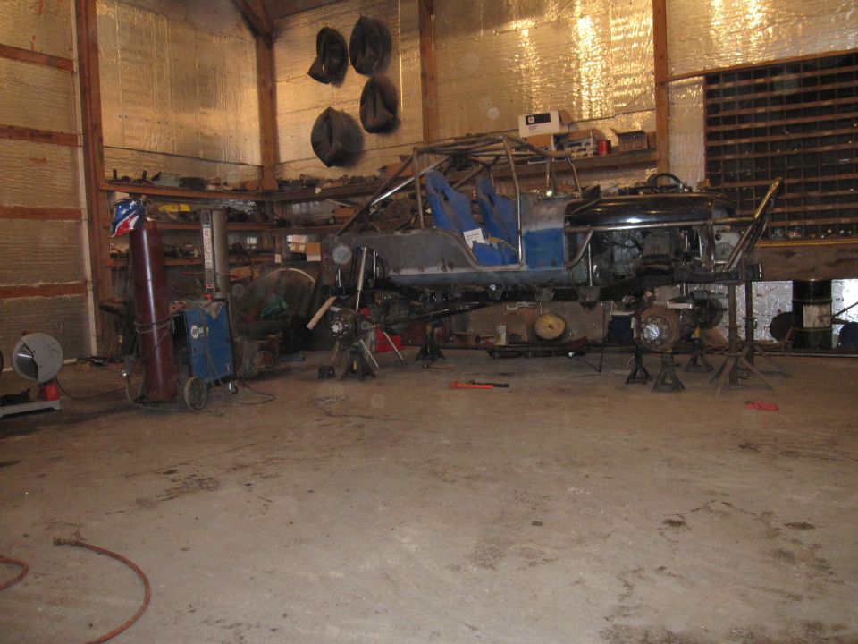 Custom buggy frame with roll cage and blue seats, raised on jack stands in a workshop