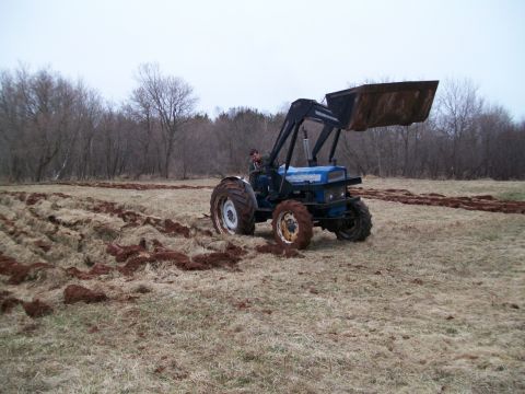 2000 Chevrolet Tahoe - A blue tractor with a front loader is actively plowing a field under an overcast sky.