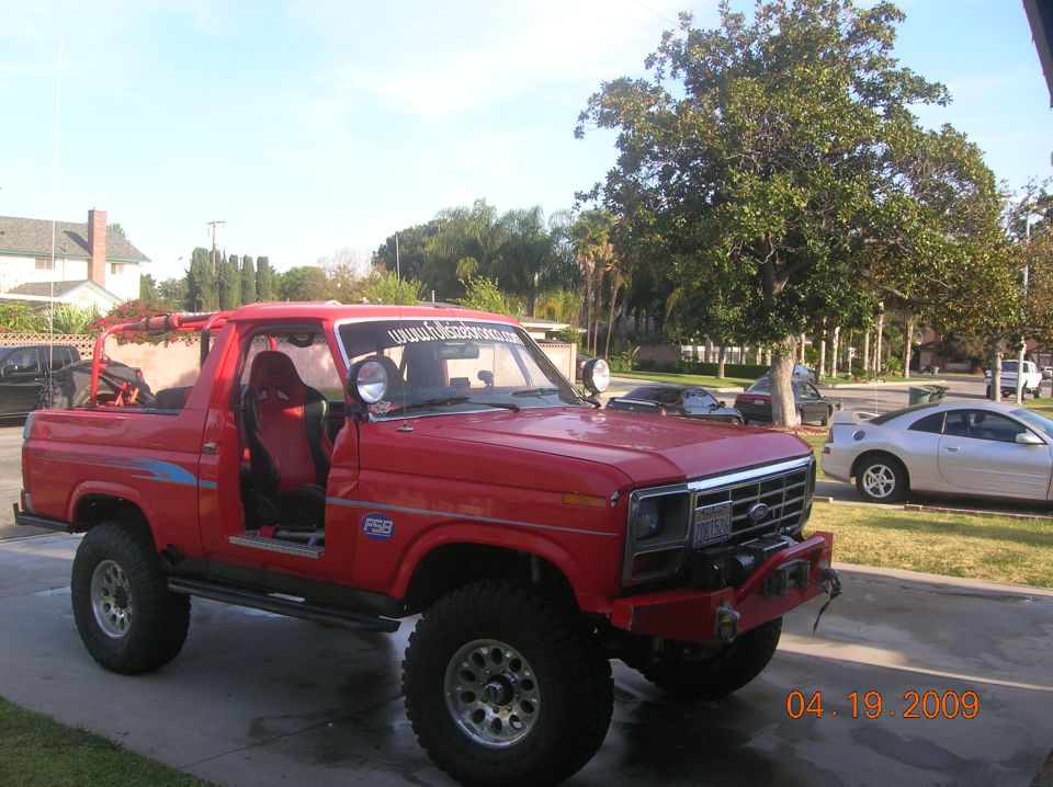 Modified red Ford Bronco with racing seats and off-road tires parked in a driveway. Another car is visible in the background