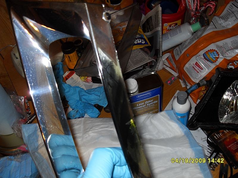 Chrome car part held by gloved hand, with mineral spirits and other cleaning supplies in background