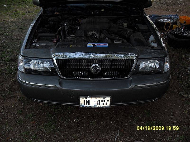 Front view of a 2004 Mercury Grand Marquis with hood open showing engine bay; tools and a spare tire are visible to the right