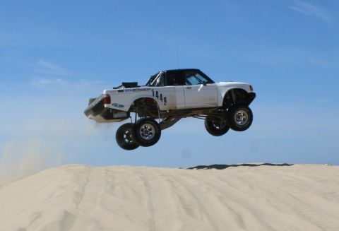 1984 Ford Bronco - A white off-road truck soars through the air over a sand dune, displaying the number 1449 on its side.