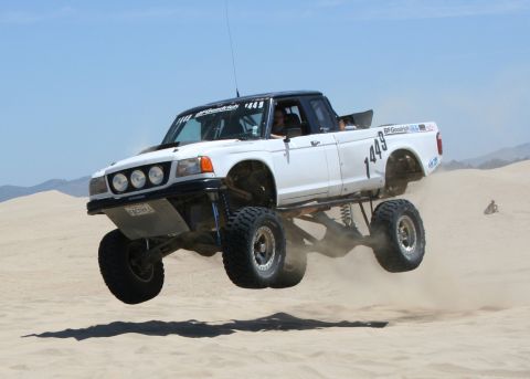 1984 Ford Bronco - This modified pickup truck is airborne over a sandy terrain, kicking up dust.