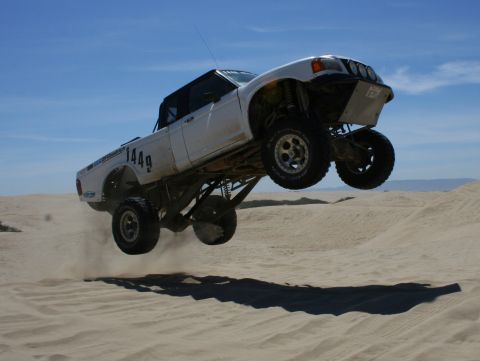 1984 Ford Bronco - A modified white truck soars through the air above a sandy desert landscape.