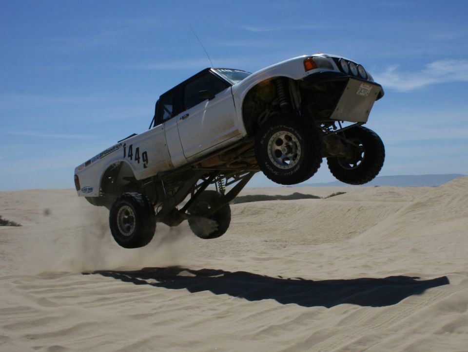 White off-road truck airborne over sand dunes, showing custom suspension and large tires