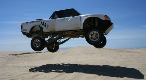 1984 Ford Bronco - This modified truck is airborne over a sandy landscape under a clear blue sky.