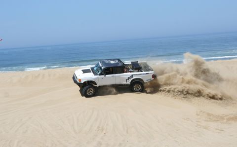 1984 Ford Bronco - A modified pickup truck navigates a sandy dune near the ocean.