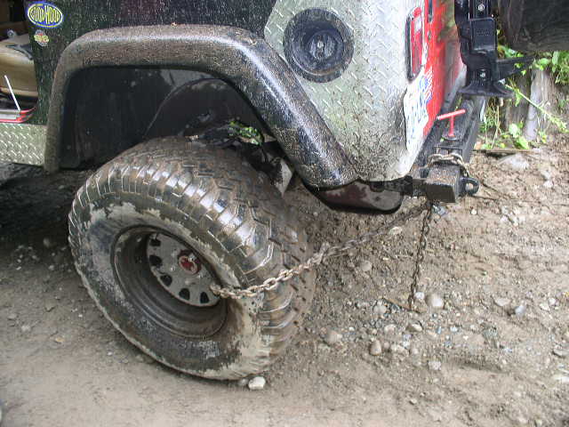Muddy rear corner of an off-road rig with knobby tire, diamond plate panel and hitch chain