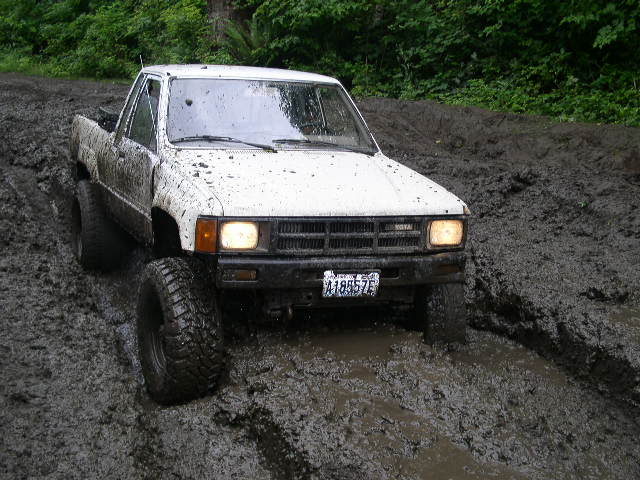 White lifted Toyota pickup covered in mud sitting in a deep mud pit, headlights on