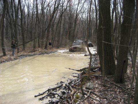 1991 Ford Bronco - Off-roading Jeeps navigate a challenging muddy water crossing in a dense forest.