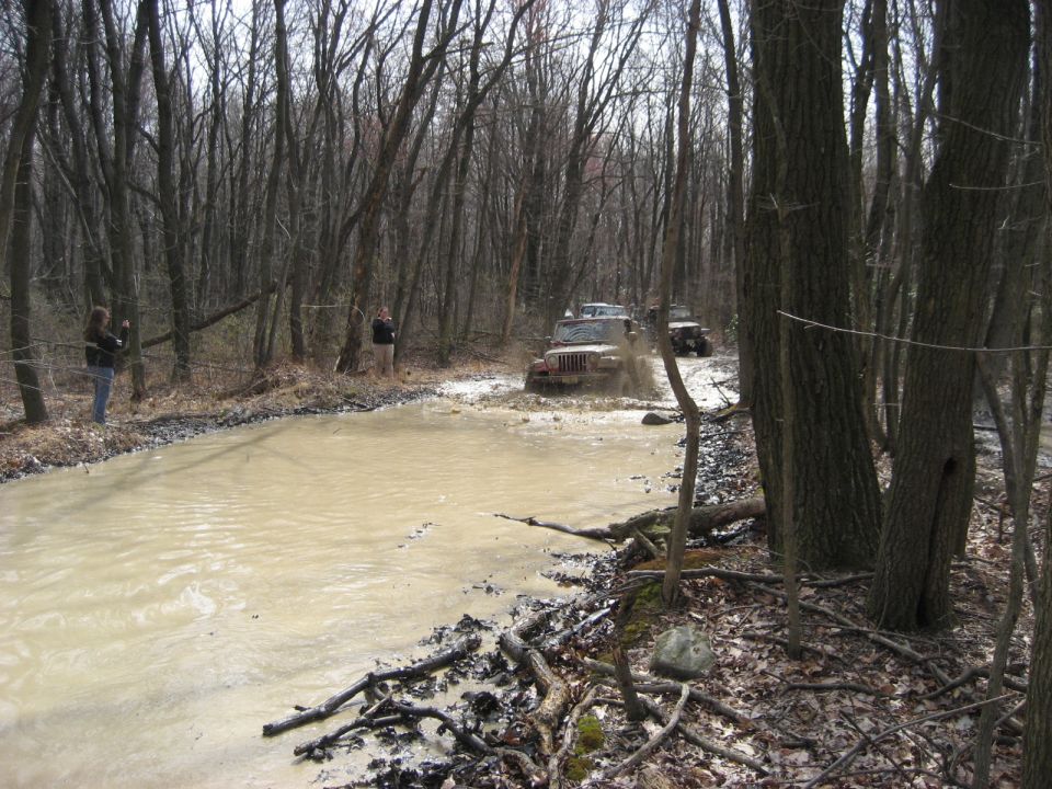 Several Jeeps driving through a muddy water obstacle on a wooded trail. Two people watch from the side