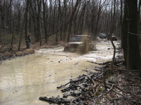 1991 Ford Bronco - Several off-road vehicles navigate a challenging muddy trail in a wooded area.