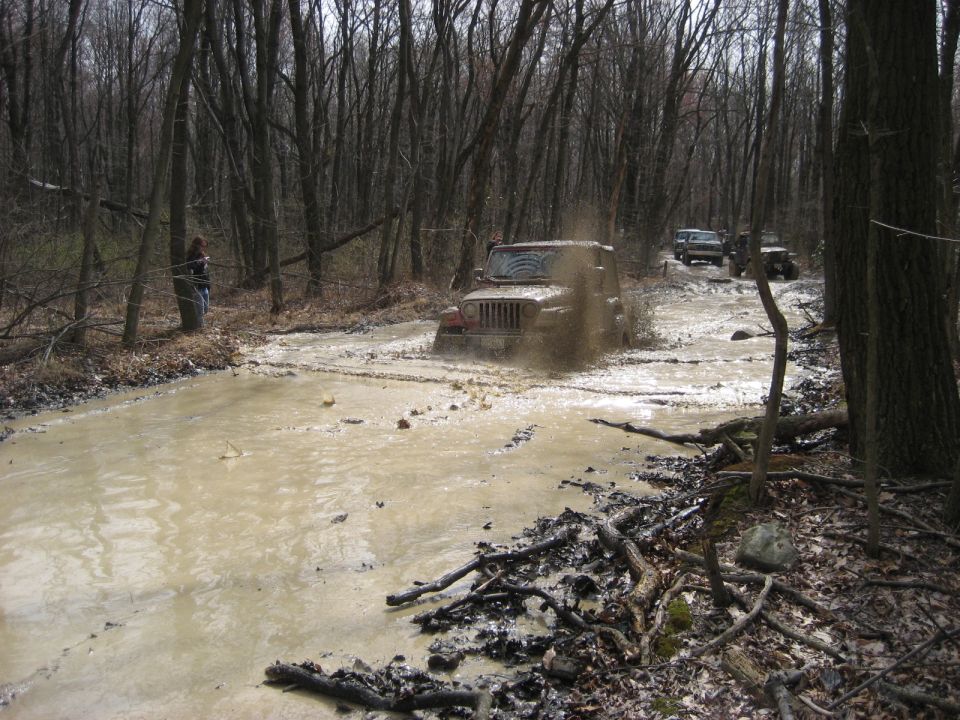 Mud-splattered Jeep Wrangler driving through a muddy forest road, kicking up water
