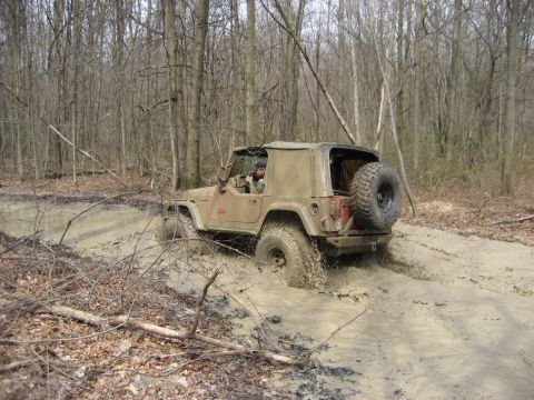 1991 Ford Bronco - Jeep Wrangler navigates a challenging muddy path in a wooded area.
