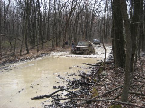 1991 Ford Bronco - Off-road vehicles navigate a challenging muddy path surrounded by bare trees.