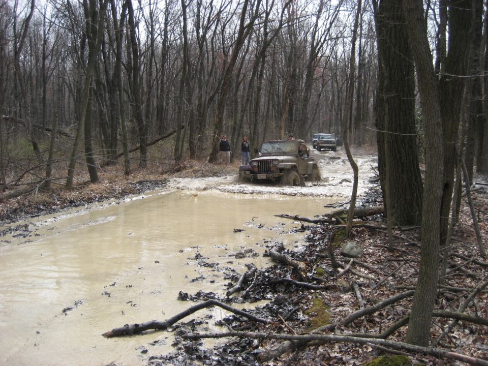 Brown Jeep Wrangler driving through a muddy forest trail with another vehicle behind it