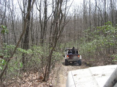 1991 Ford Bronco - Two off-road vehicles navigate a wooded path with loose rocks under a cloudy sky.