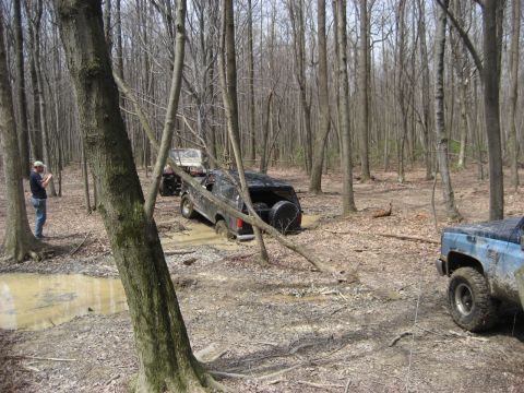 1991 Ford Bronco - Multiple off-road vehicles navigate a muddy forest trail, with one Bronco deeply mired in the muck.