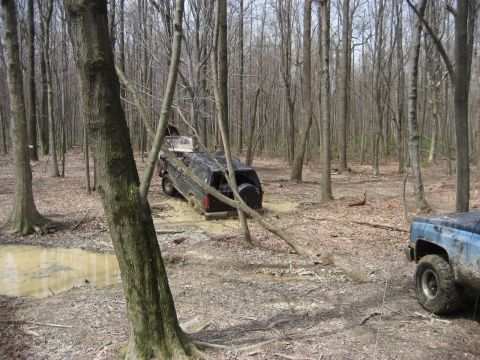 1991 Ford Bronco - Two vehicles navigate a muddy, wooded area, with one SUV deeply stuck in the mire.