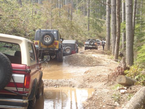 1989 Ford Bronco - A group of off-road vehicles navigate a challenging muddy path through a dense forest.