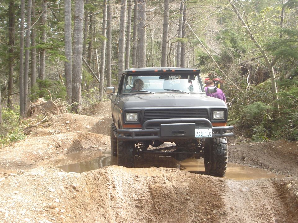 Dark gray Ford Bronco driving through muddy terrain with trees in the background